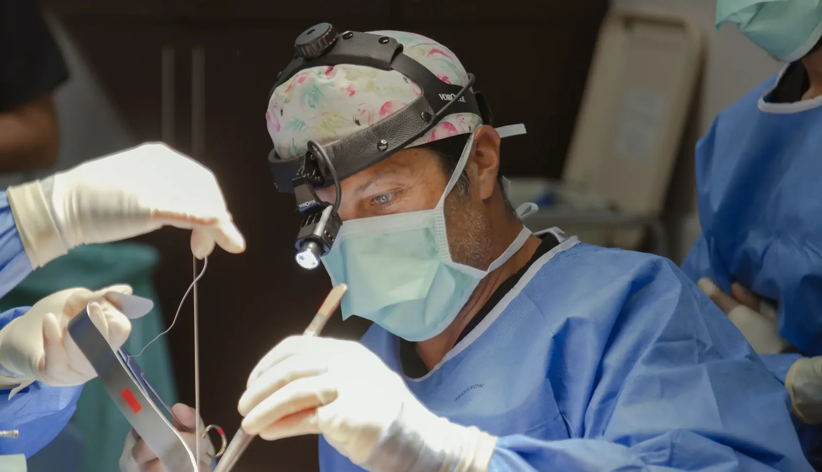 A male patient lying shirtless on a treatment bed wearing a disposable hair net, while a medical professional in a white lab coat and blue latex gloves applies a body sculpting device to his abdomen in a clinical setting.