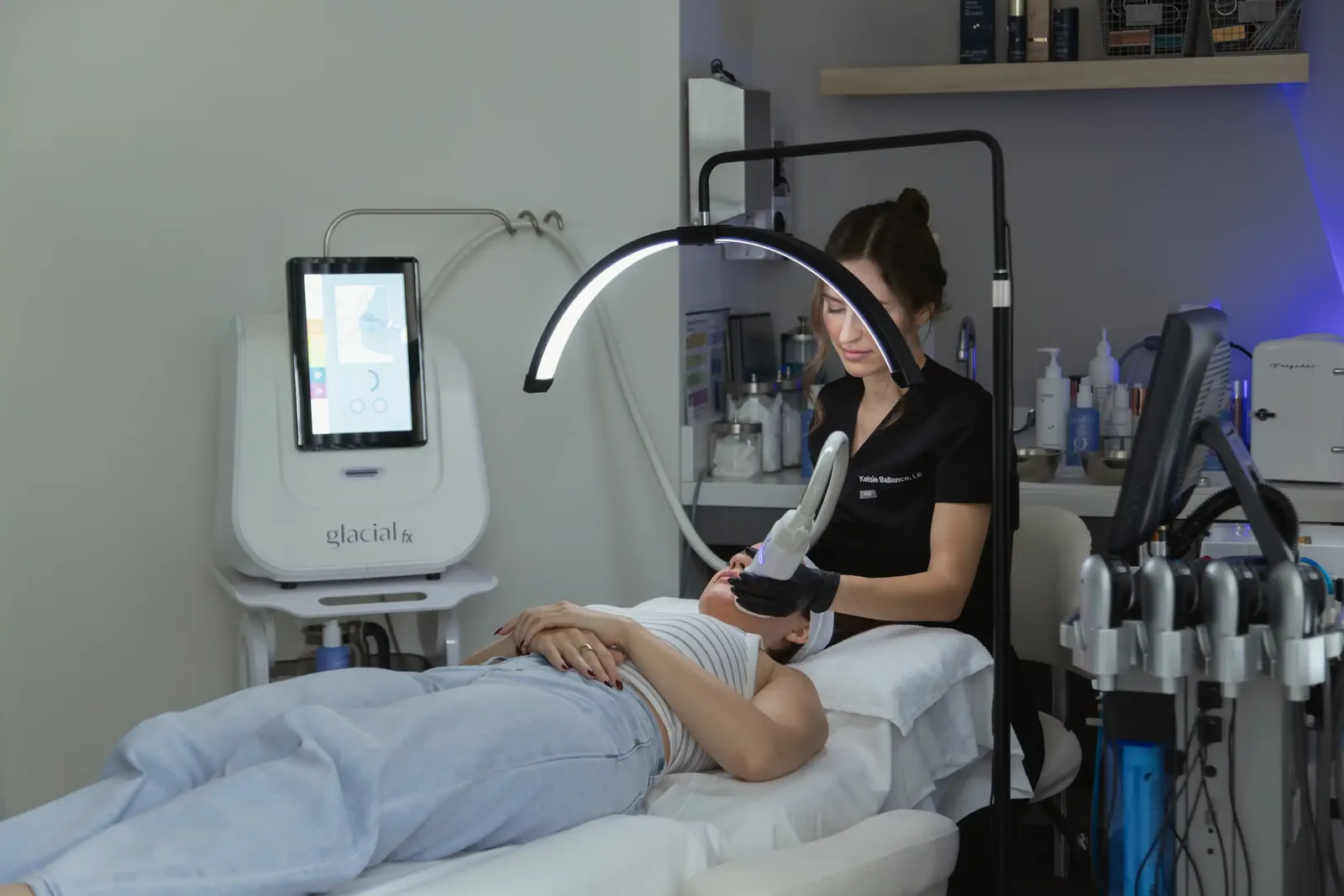A male patient lying shirtless on a treatment bed wearing a disposable hair net, while a medical professional in a white lab coat and blue latex gloves applies a body sculpting device to his abdomen in a clinical setting.