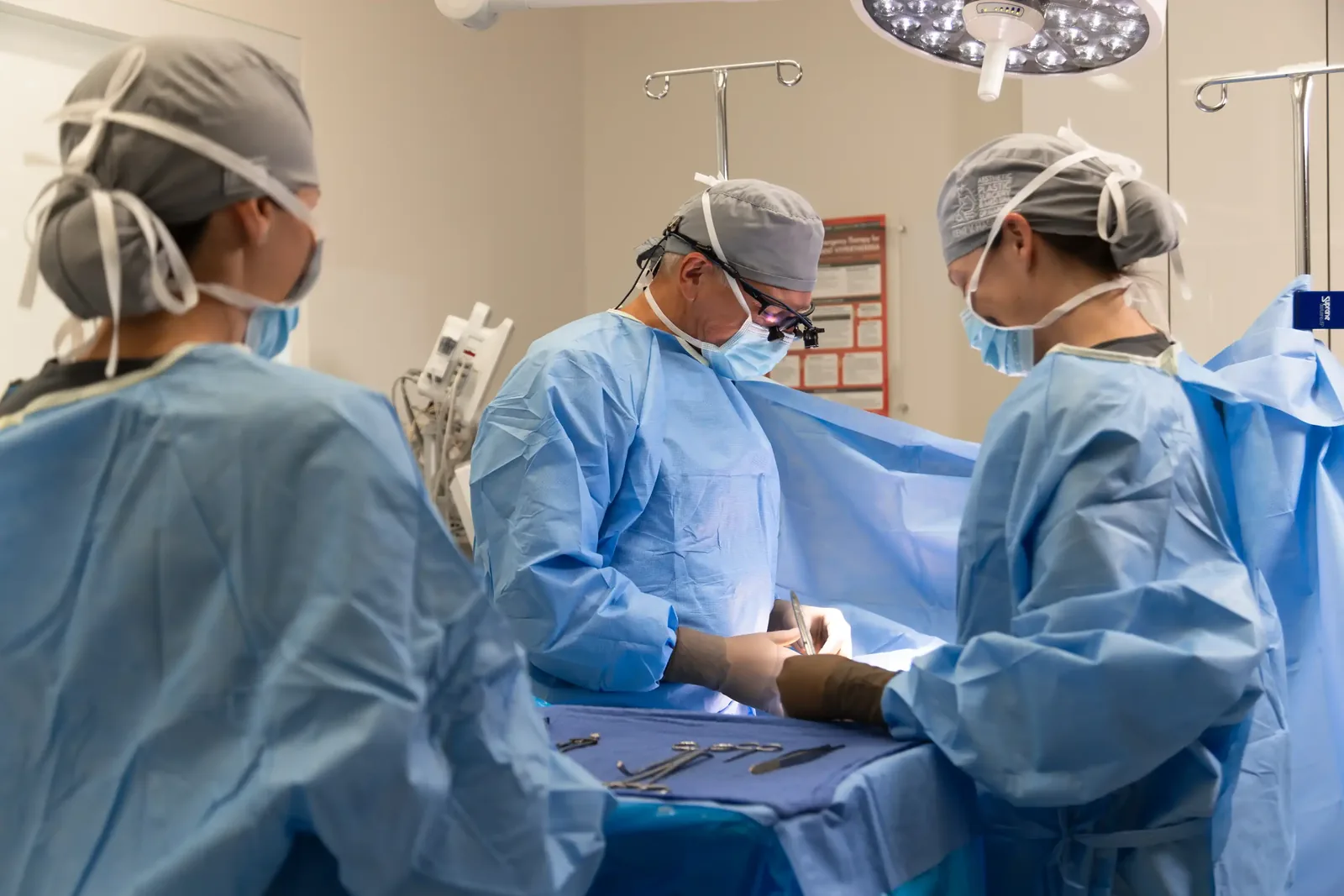 A male patient lying shirtless on a treatment bed wearing a disposable hair net, while a medical professional in a white lab coat and blue latex gloves applies a body sculpting device to his abdomen in a clinical setting.