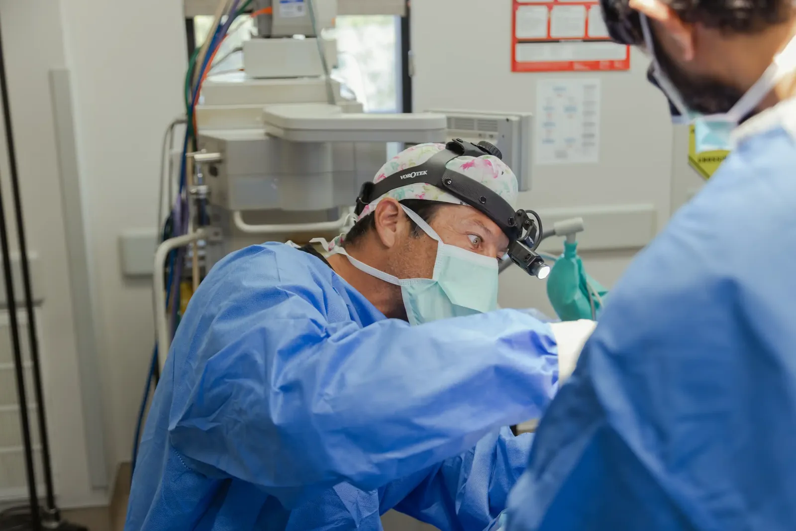 A male patient lying shirtless on a treatment bed wearing a disposable hair net, while a medical professional in a white lab coat and blue latex gloves applies a body sculpting device to his abdomen in a clinical setting.