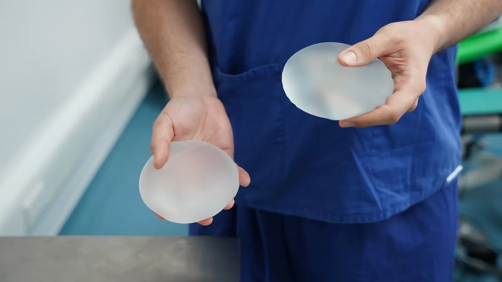 A male patient lying shirtless on a treatment bed wearing a disposable hair net, while a medical professional in a white lab coat and blue latex gloves applies a body sculpting device to his abdomen in a clinical setting.