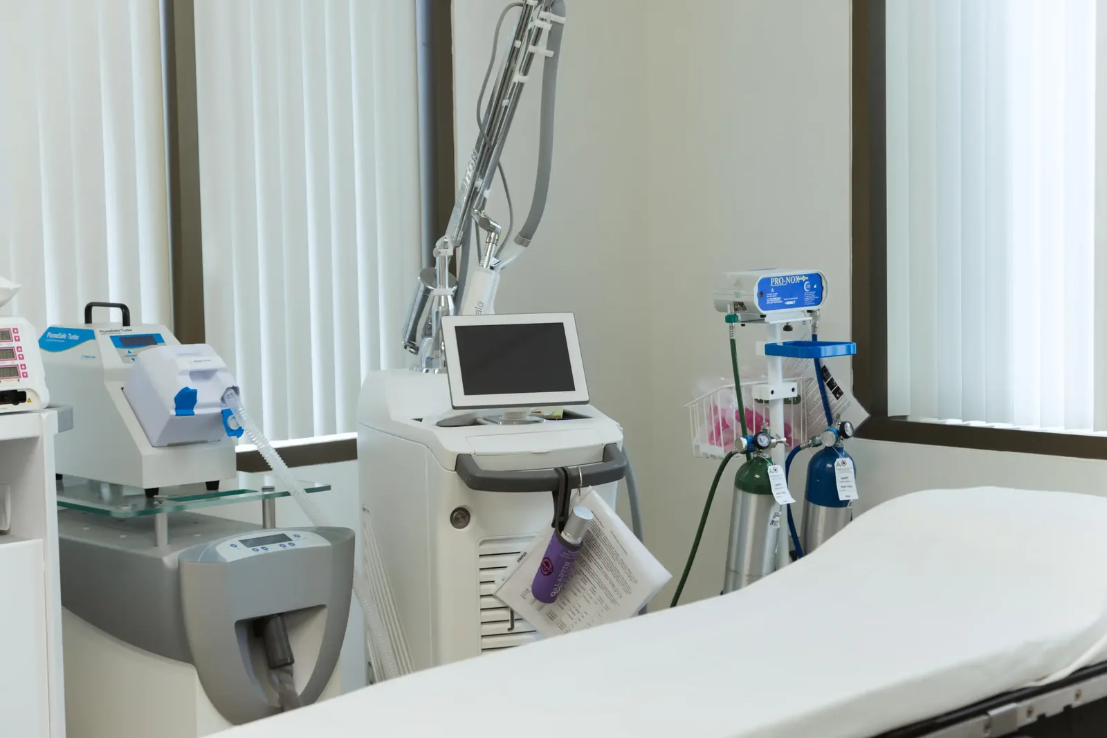 A male patient lying shirtless on a treatment bed wearing a disposable hair net, while a medical professional in a white lab coat and blue latex gloves applies a body sculpting device to his abdomen in a clinical setting.