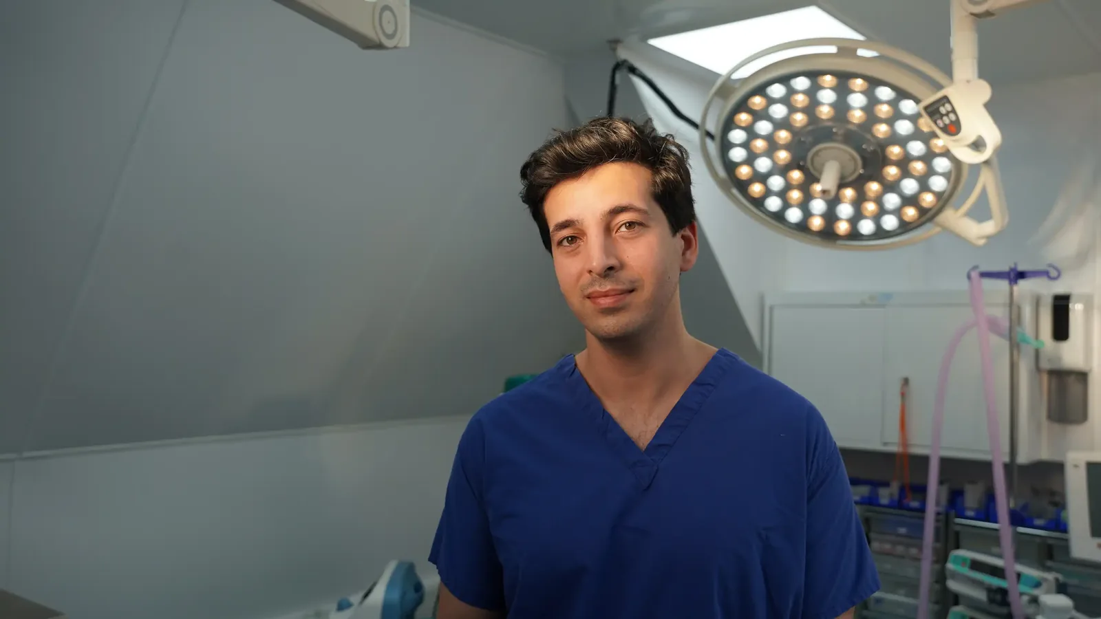 A male patient lying shirtless on a treatment bed wearing a disposable hair net, while a medical professional in a white lab coat and blue latex gloves applies a body sculpting device to his abdomen in a clinical setting.