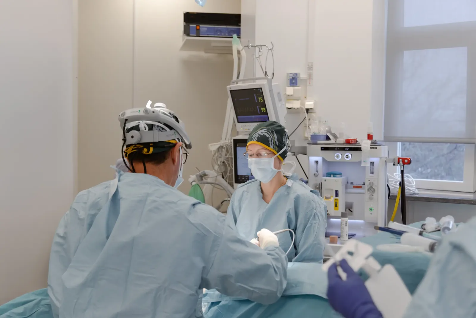 A male patient lying shirtless on a treatment bed wearing a disposable hair net, while a medical professional in a white lab coat and blue latex gloves applies a body sculpting device to his abdomen in a clinical setting.