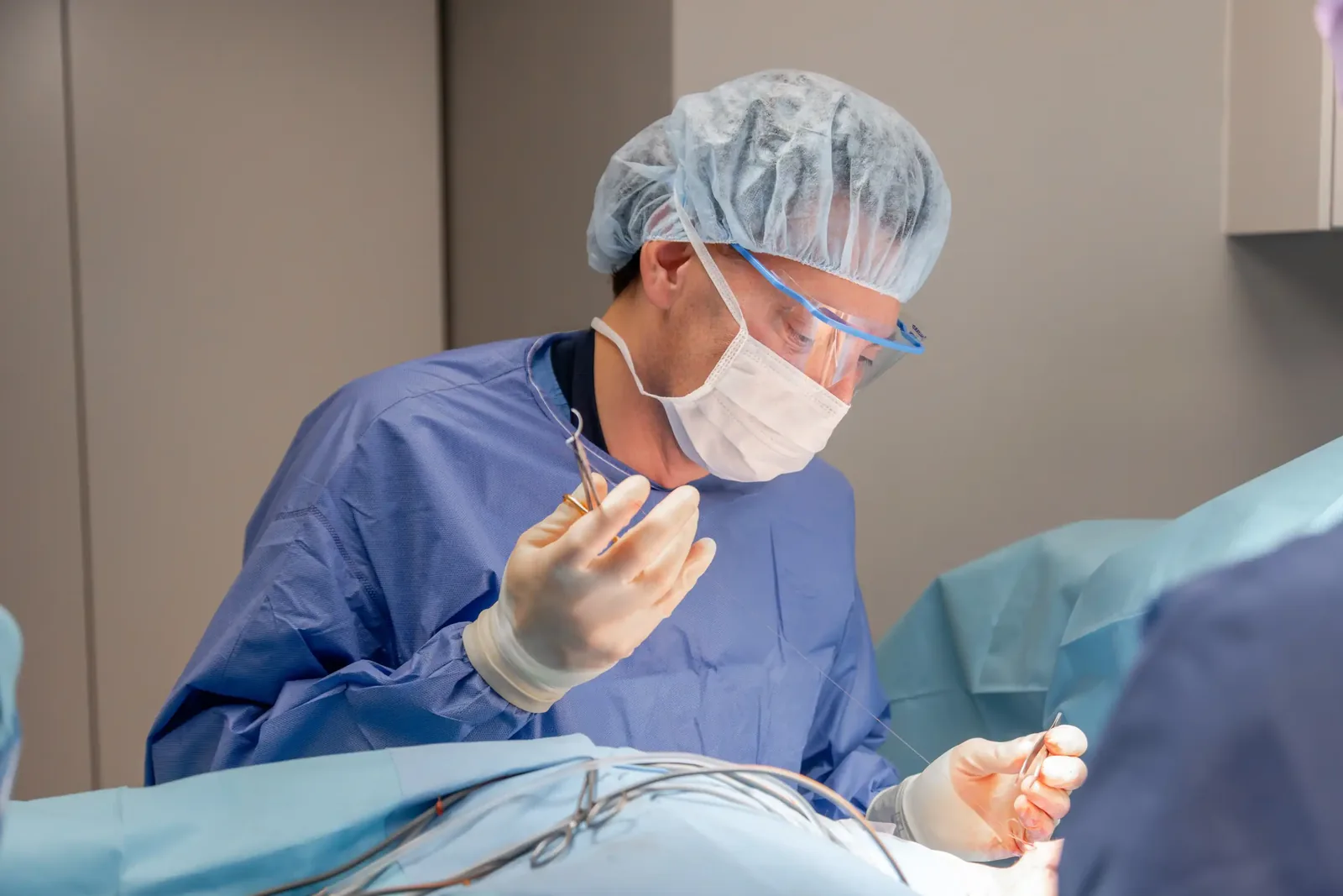 A male patient lying shirtless on a treatment bed wearing a disposable hair net, while a medical professional in a white lab coat and blue latex gloves applies a body sculpting device to his abdomen in a clinical setting.