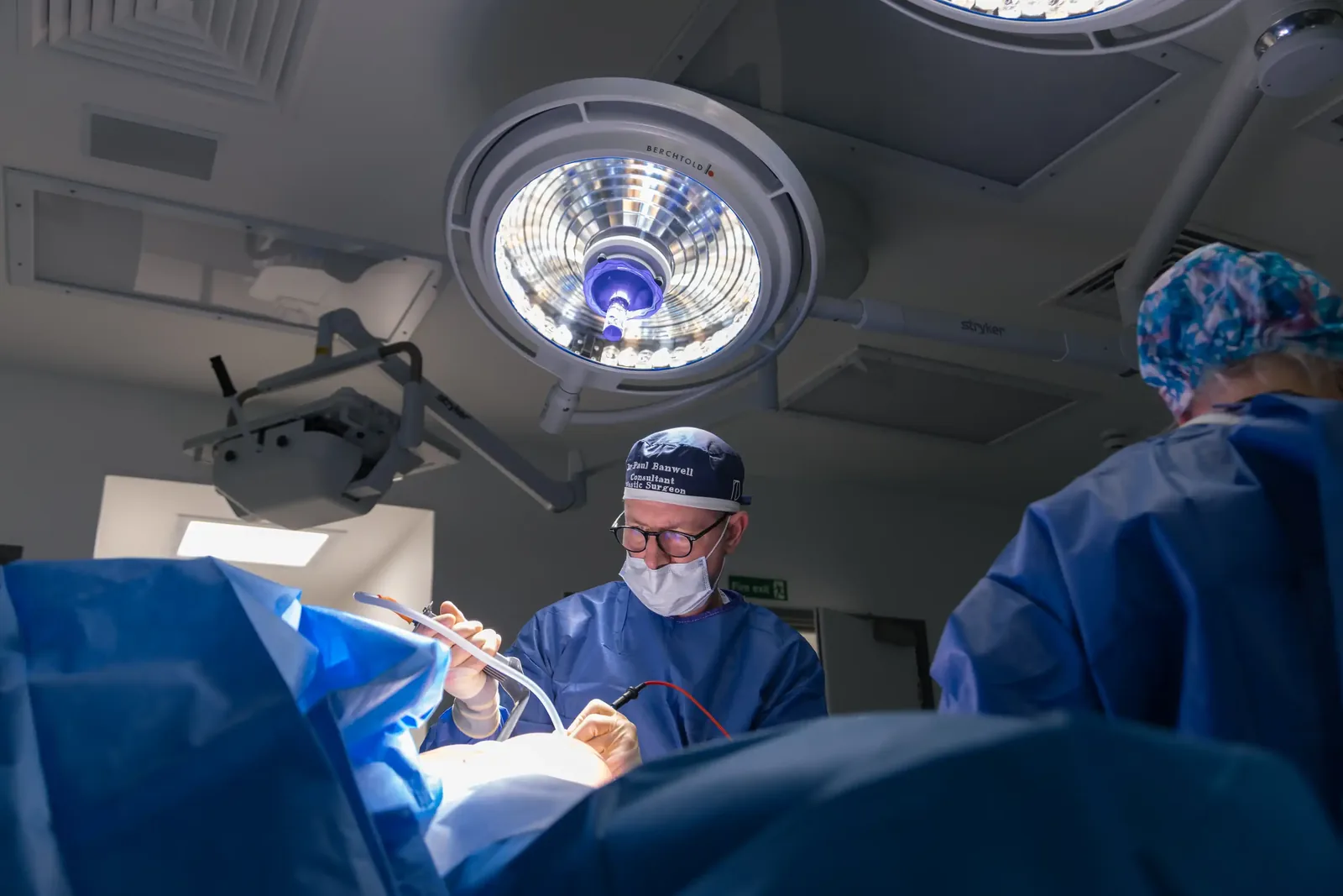 A male patient lying shirtless on a treatment bed wearing a disposable hair net, while a medical professional in a white lab coat and blue latex gloves applies a body sculpting device to his abdomen in a clinical setting.