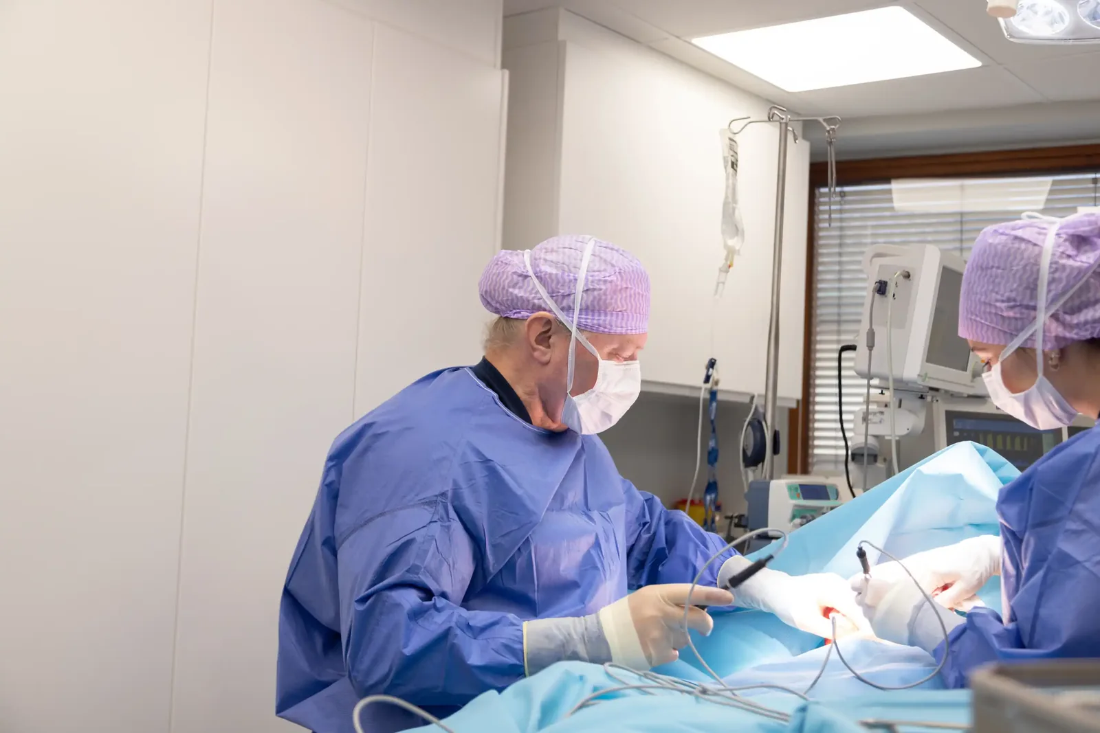 A male patient lying shirtless on a treatment bed wearing a disposable hair net, while a medical professional in a white lab coat and blue latex gloves applies a body sculpting device to his abdomen in a clinical setting.