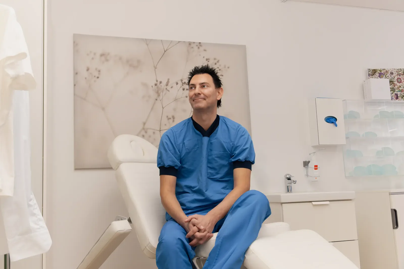 A male patient lying shirtless on a treatment bed wearing a disposable hair net, while a medical professional in a white lab coat and blue latex gloves applies a body sculpting device to his abdomen in a clinical setting.