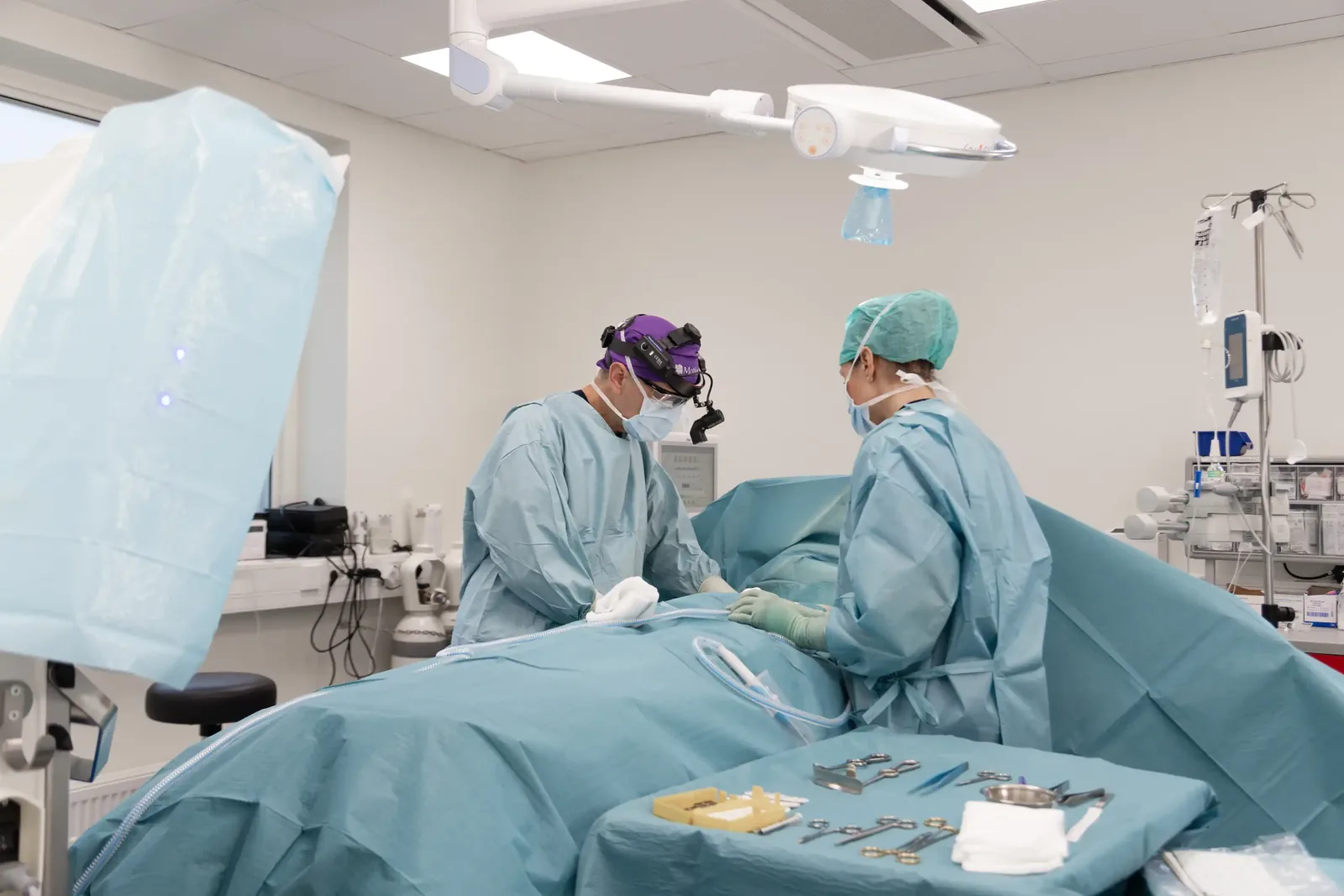 A male patient lying shirtless on a treatment bed wearing a disposable hair net, while a medical professional in a white lab coat and blue latex gloves applies a body sculpting device to his abdomen in a clinical setting.