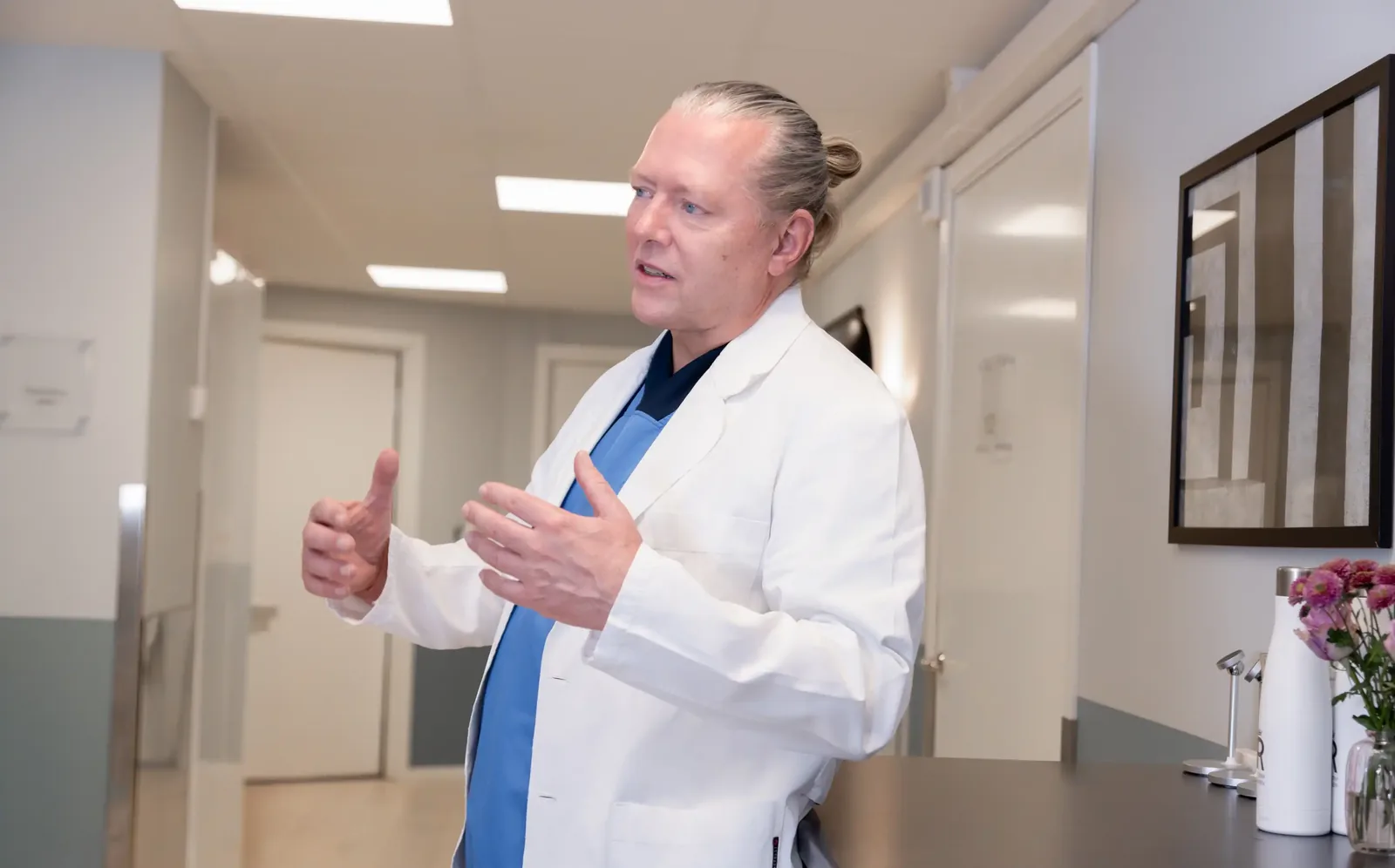 A male patient lying shirtless on a treatment bed wearing a disposable hair net, while a medical professional in a white lab coat and blue latex gloves applies a body sculpting device to his abdomen in a clinical setting.
