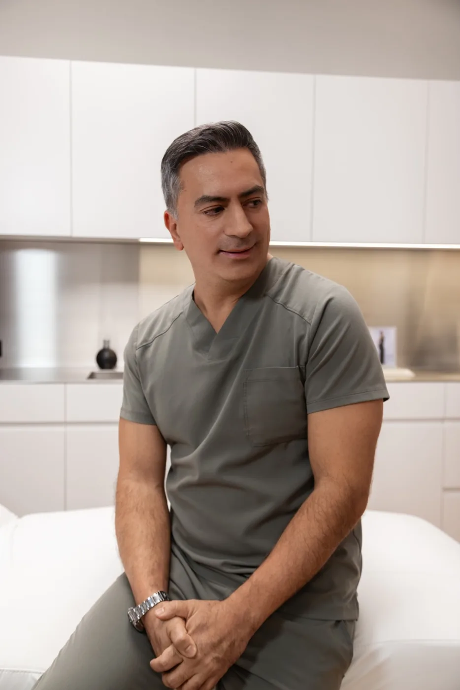 Associate Professor Dr. Farid Rezaeian, a male medical professional with short salt-and-pepper hair, seated and wearing olive-green scrubs with a chest pocket and a silver watch, in a modern clinical setting with white cabinetry.
