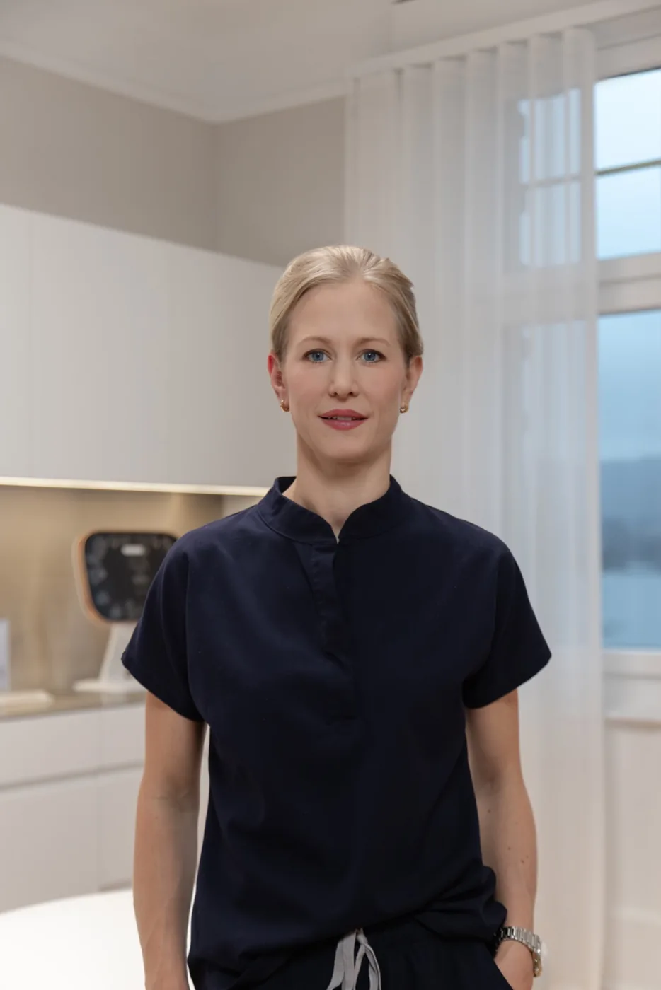Dr. Martina Schneider, a blonde woman with her hair pulled back, wearing a navy blue short-sleeve medical scrub top and a silver watch, standing in a clean, modern clinical or medical office setting.