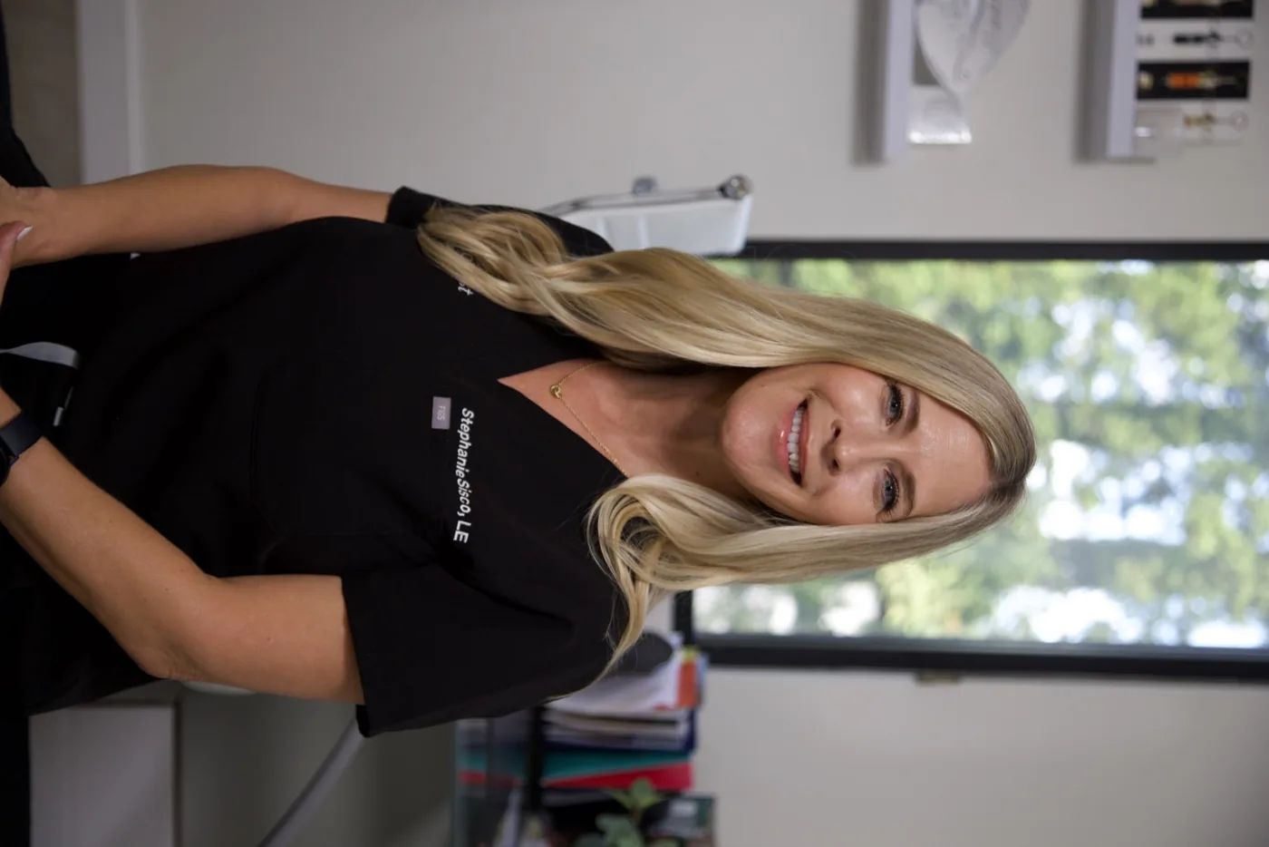Professional headshot of Stephanie Sisco, LE, a blonde woman smiling and wearing black scrubs with her name embroidered on the chest, photographed in a clinical or medical office setting.