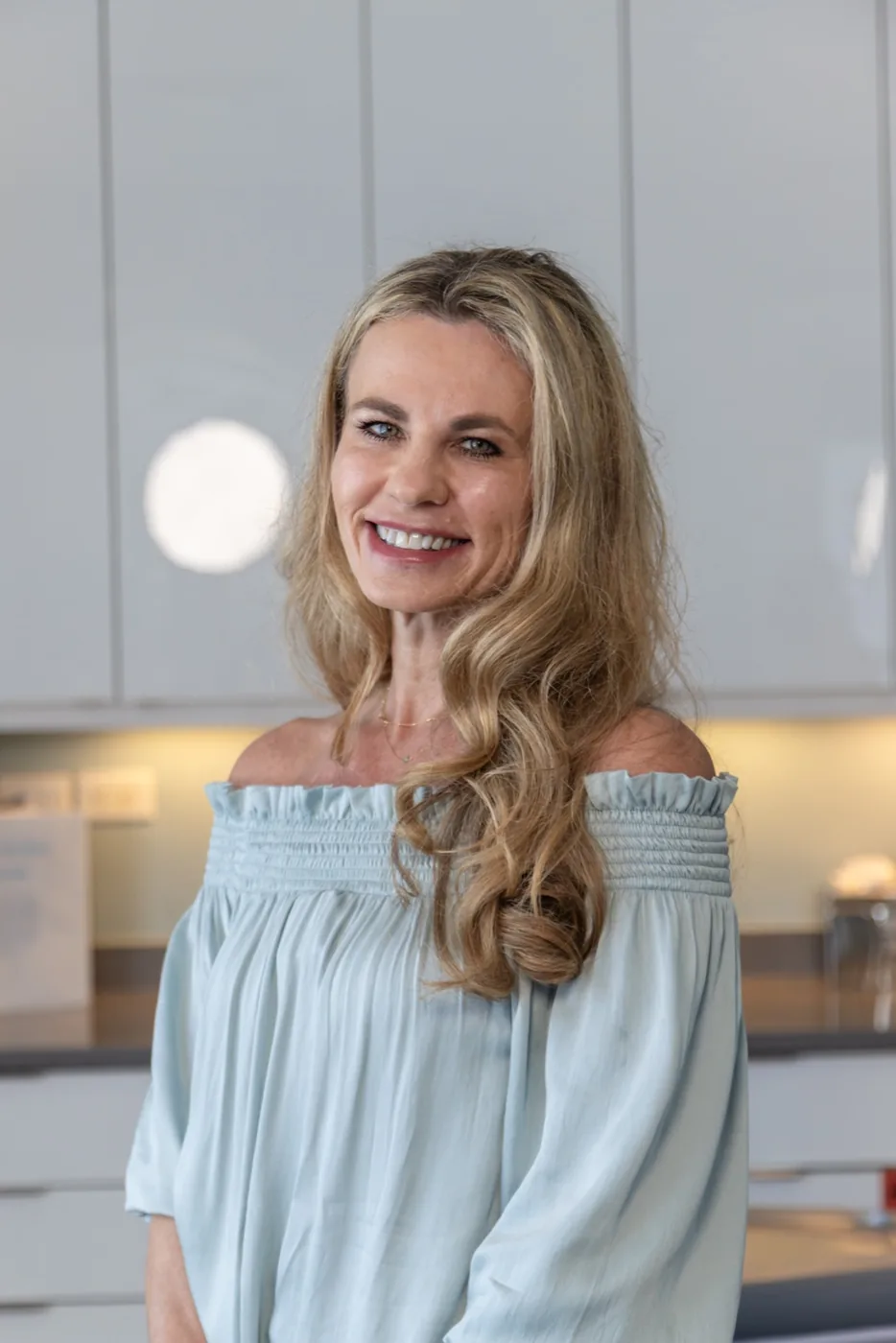 Amanda Walczak, RN — a smiling woman with long wavy blonde hair wearing a light blue off-the-shoulder top, photographed in a bright modern kitchen setting.
