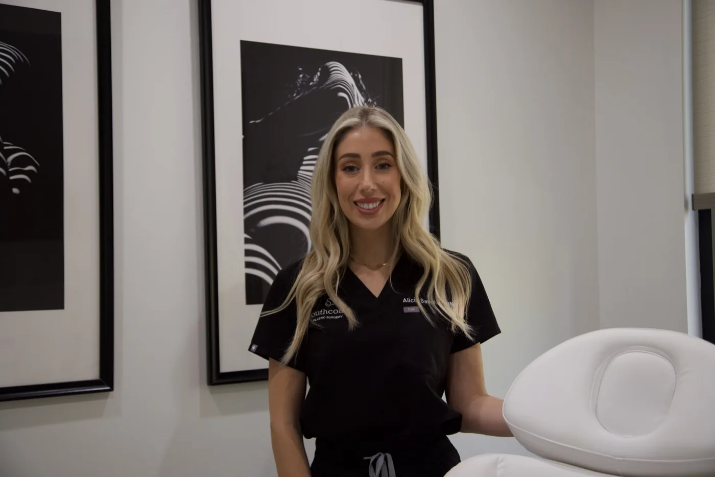 Alicia Sanchez, RN, smiling and standing in a clinic room wearing a black medical scrub top with a logo and name badge. Behind her are two framed black-and-white abstract art prints on a white wall, and to her right is a white treatment chair.