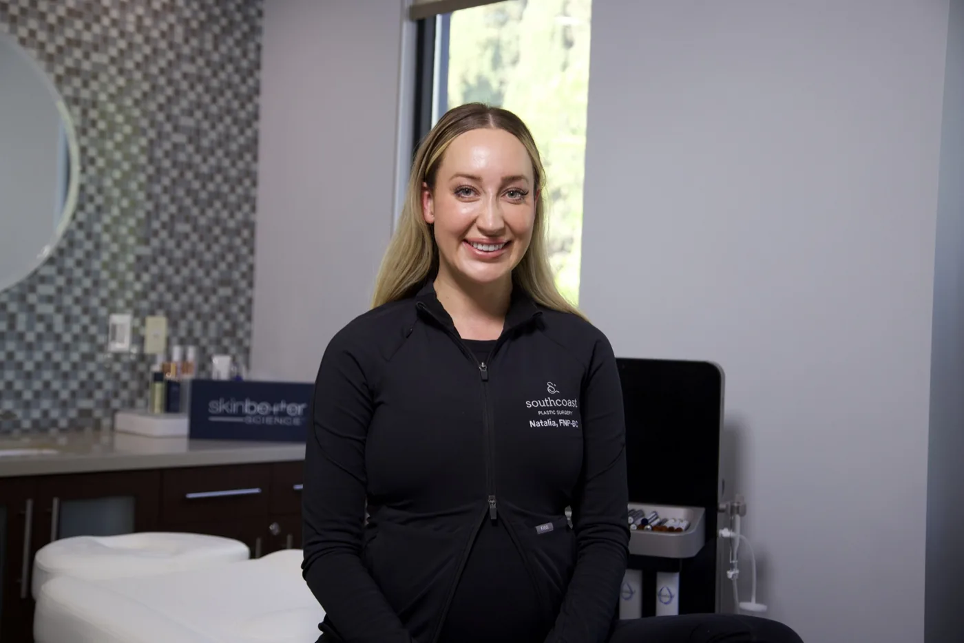 Natalia Srabian, RN, a healthcare professional at South Coast Plastic Surgery, smiling and posing in a medical treatment room wearing a black zip-up jacket embroidered with the South Coast logo and her name and credentials. Skinbetter Science products and