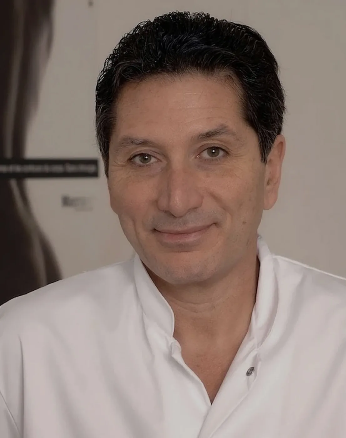 Portrait of Dr. Foued Hamza, a middle-aged man with short dark hair and hazel eyes, smiling warmly, wearing a white medical coat or scrub top, with a softly blurred indoor background.