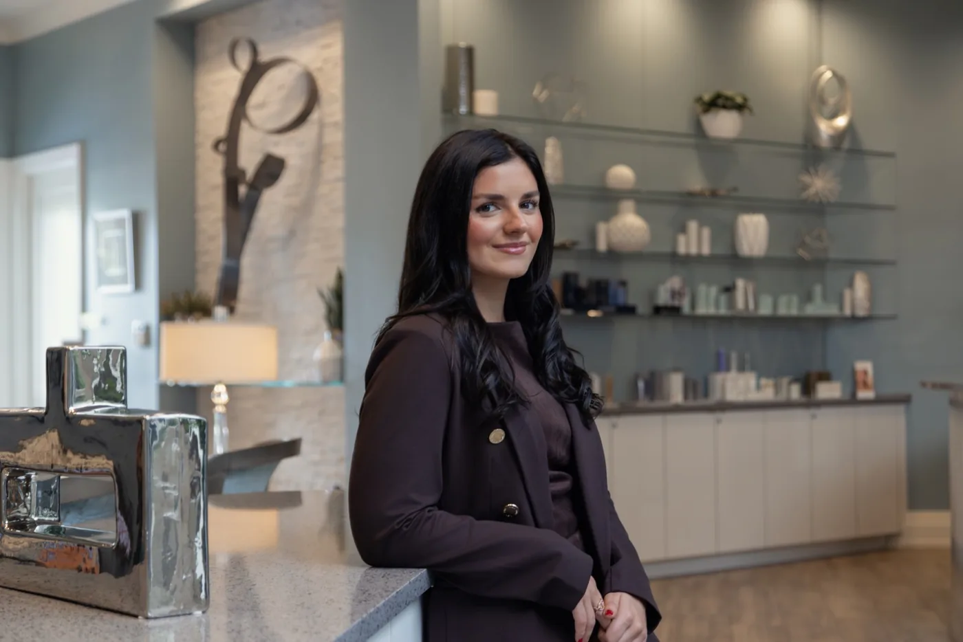 Daniela Valdes, FNP-C, a woman with long dark hair wearing a dark burgundy blazer, smiling and leaning against a counter inside a modern medical spa or aesthetic clinic, with shelved skincare products and decorative items visible in the background.