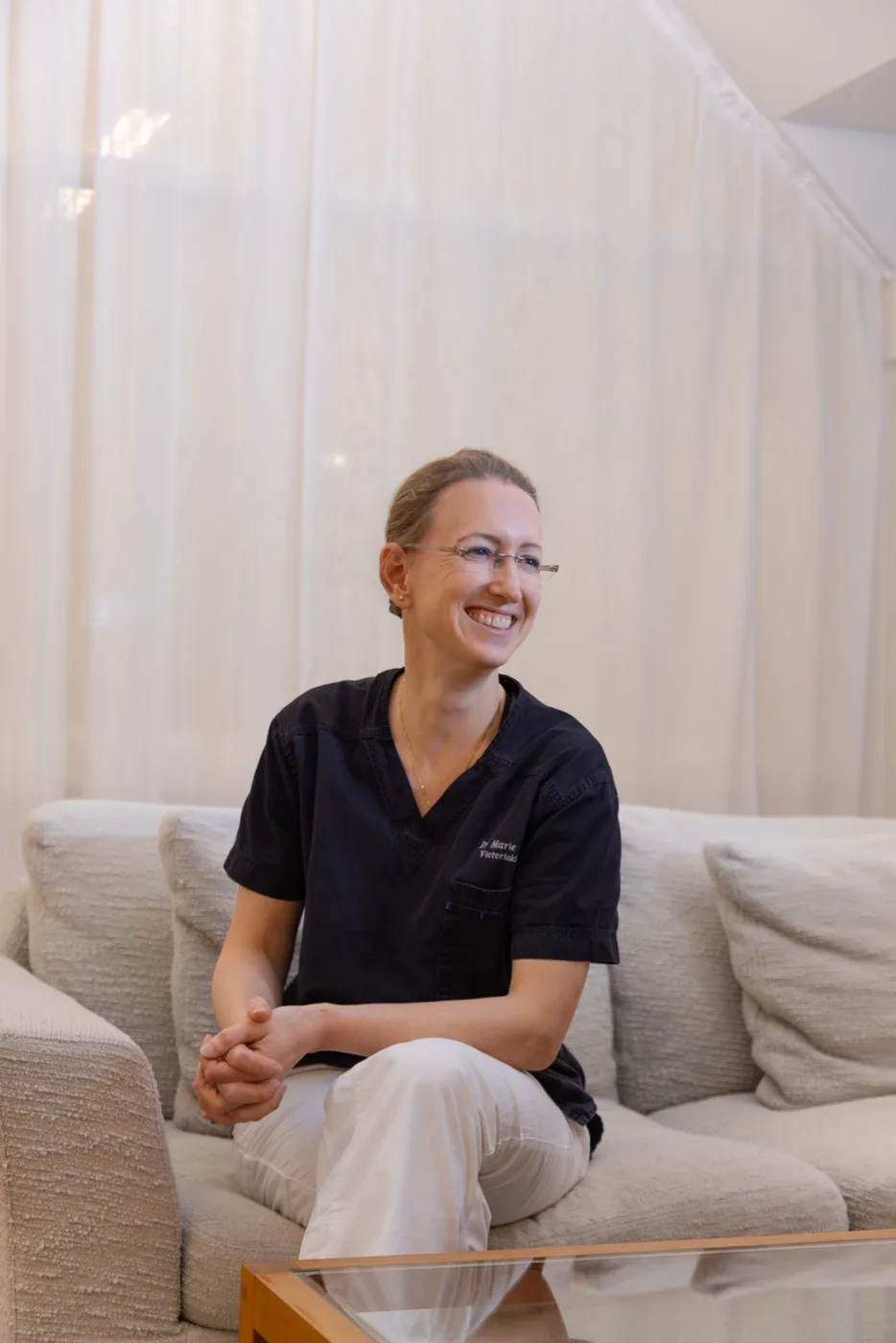 Dr. Marie Jaeger, a smiling woman wearing glasses and dark navy medical scrubs with a name badge, sitting relaxed on a light gray sofa with her hands clasped, in a bright, airy room with white curtains in the background.