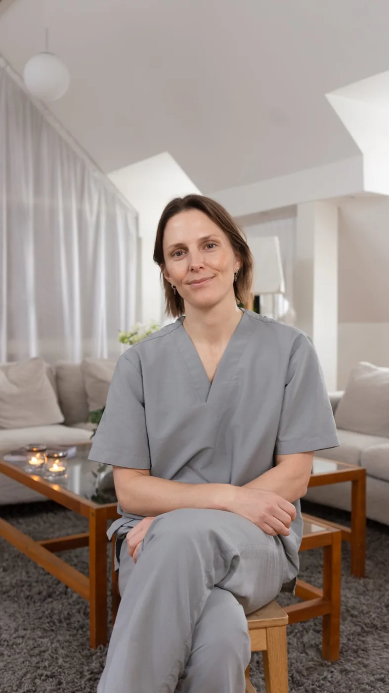 Dr. Stina Rittri, a woman with short dark hair and drop earrings, sits relaxed with arms crossed and legs crossed, wearing gray medical scrubs, in a bright, minimalist living room setting with a glass coffee table, lit tea candles, and white sofas in the