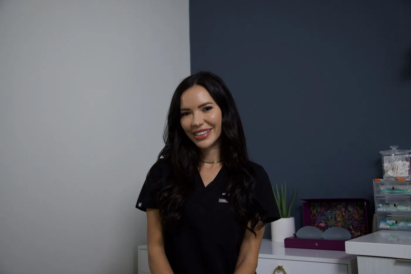 Megan Atchison, RN, BSN, a healthcare professional with long dark wavy hair, smiling warmly while wearing black medical scrubs and a gold necklace, seated in a clinical setting with medical supply containers and a small plant visible in the background.
