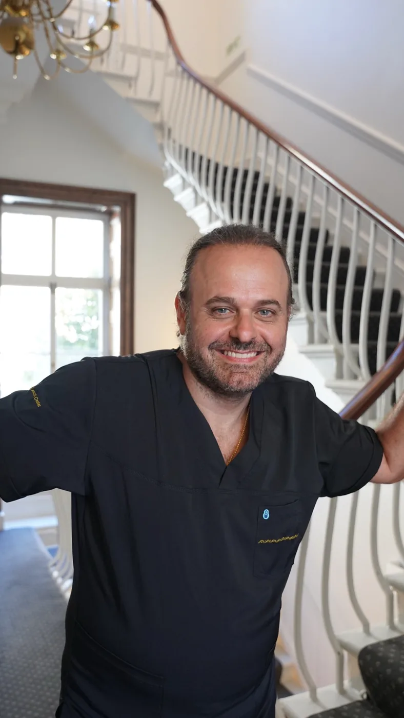 Professor Marcos Sforza, a smiling man with dark hair and a beard, wearing black medical scrubs, posing confidently in front of an elegant curved staircase with white balusters and a wooden handrail inside a bright interior space.