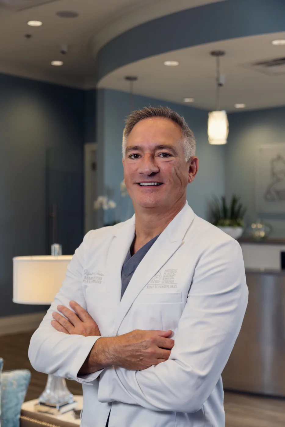 Dr. Kent V. Hasen, MD, a middle-aged man with short salt-and-pepper hair, smiling with arms crossed, wearing a white medical coat over a gray scrub top, standing in a modern, softly lit medical office reception area.