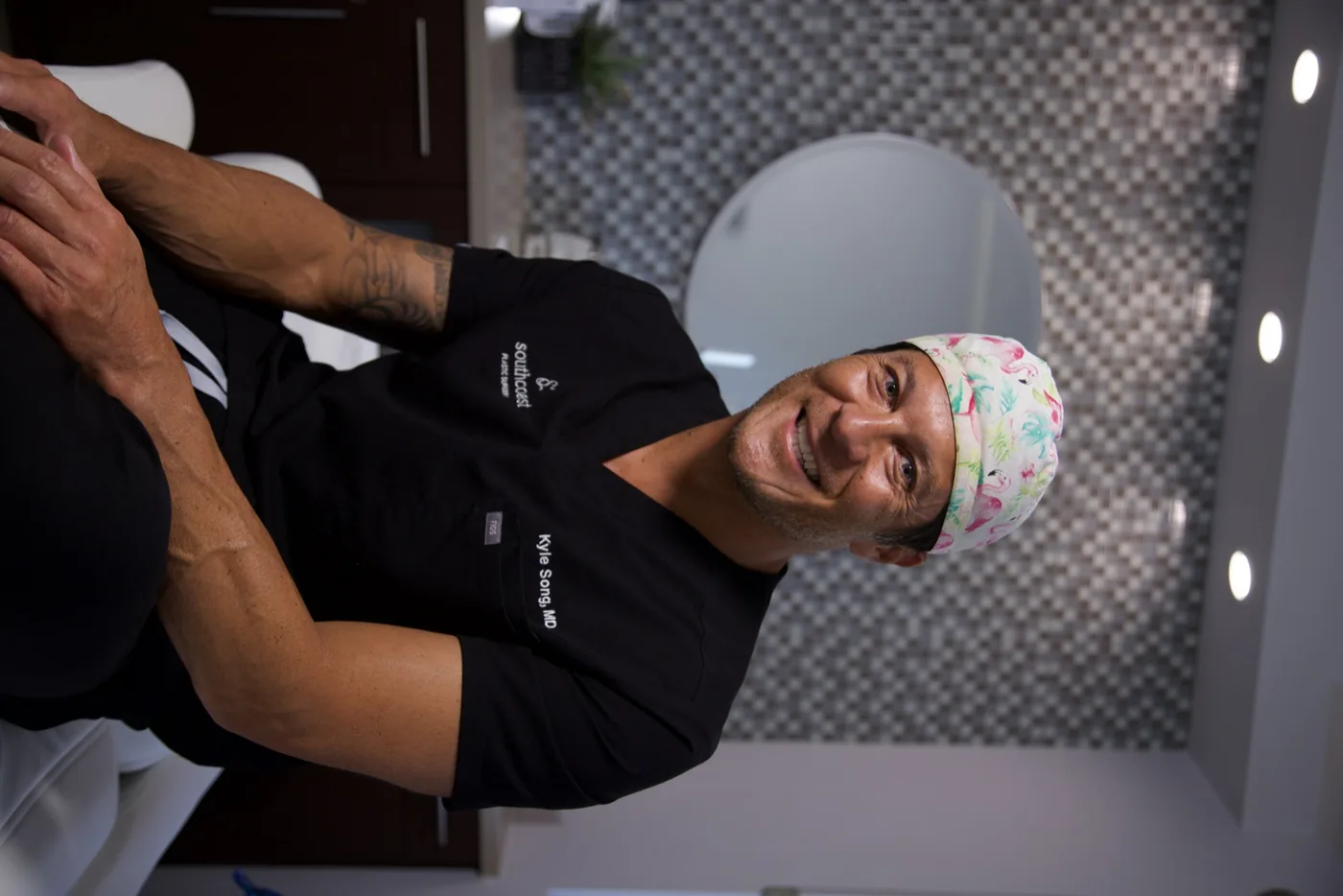 Dr. Kyle Song, MD, of Southcoast Plastic Surgery, smiling and seated in a clinical office setting. He is wearing black medical scrubs embroidered with 'Southcoast Plastic Surgery' and 'Kyle Song, MD,' along with a colorful surgical cap featuring a