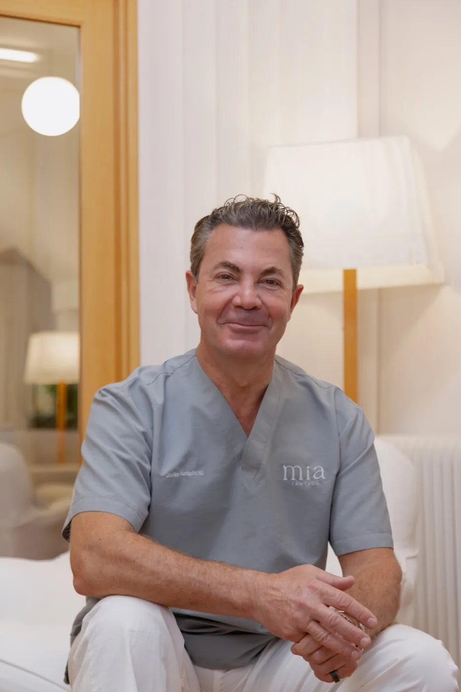 Dr. Charles Randquist, a smiling middle-aged man with dark salt-and-pepper hair, seated and wearing gray medical scrubs embroidered with the 'mia' clinic logo, in a bright, warmly lit clinical room with a floor lamp and mirror in the background.