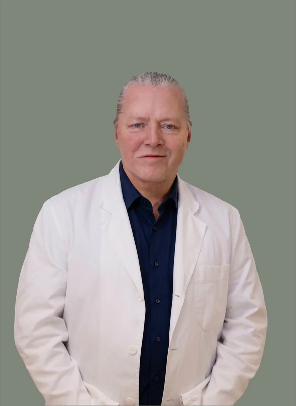 Professional portrait of Dr. Stina Klasson, a middle-aged man with slicked-back salt-and-pepper hair, wearing a white medical lab coat over a dark navy button-up shirt, posed with hands in pockets and looking directly at the camera.