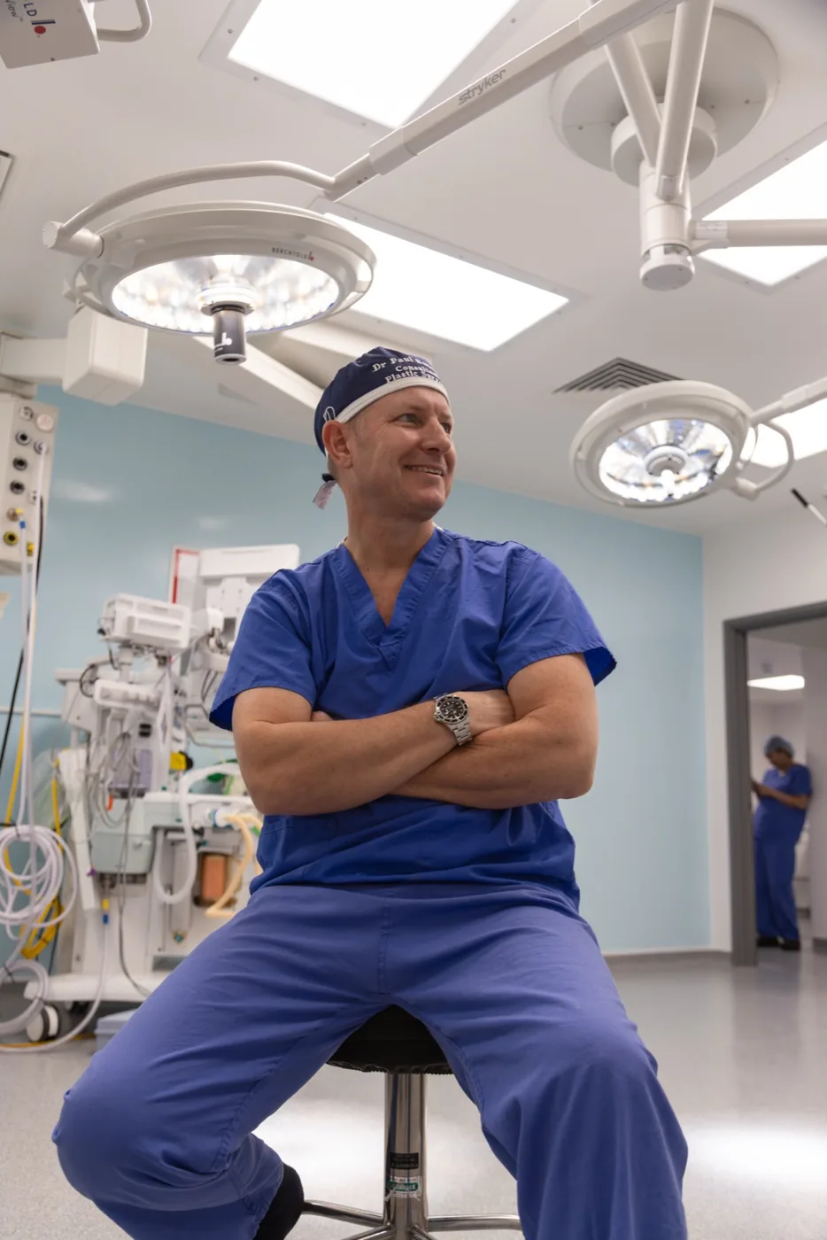 Mr. Paul Banwell, a male surgeon wearing blue scrubs and a surgical cap, sits on a stool with arms crossed and a smile, inside a modern operating theatre.