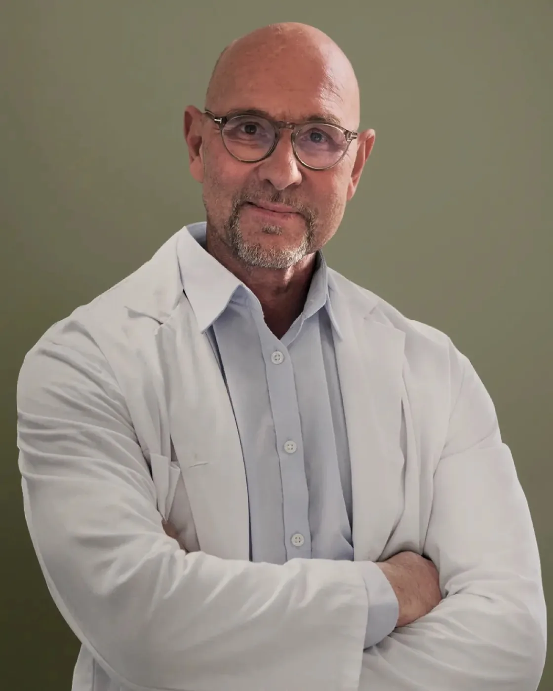 Dr. Peter Cosmo, a bald middle-aged man wearing round glasses, a white medical lab coat, and a light blue button-up shirt, standing with his arms crossed and looking directly at the camera.