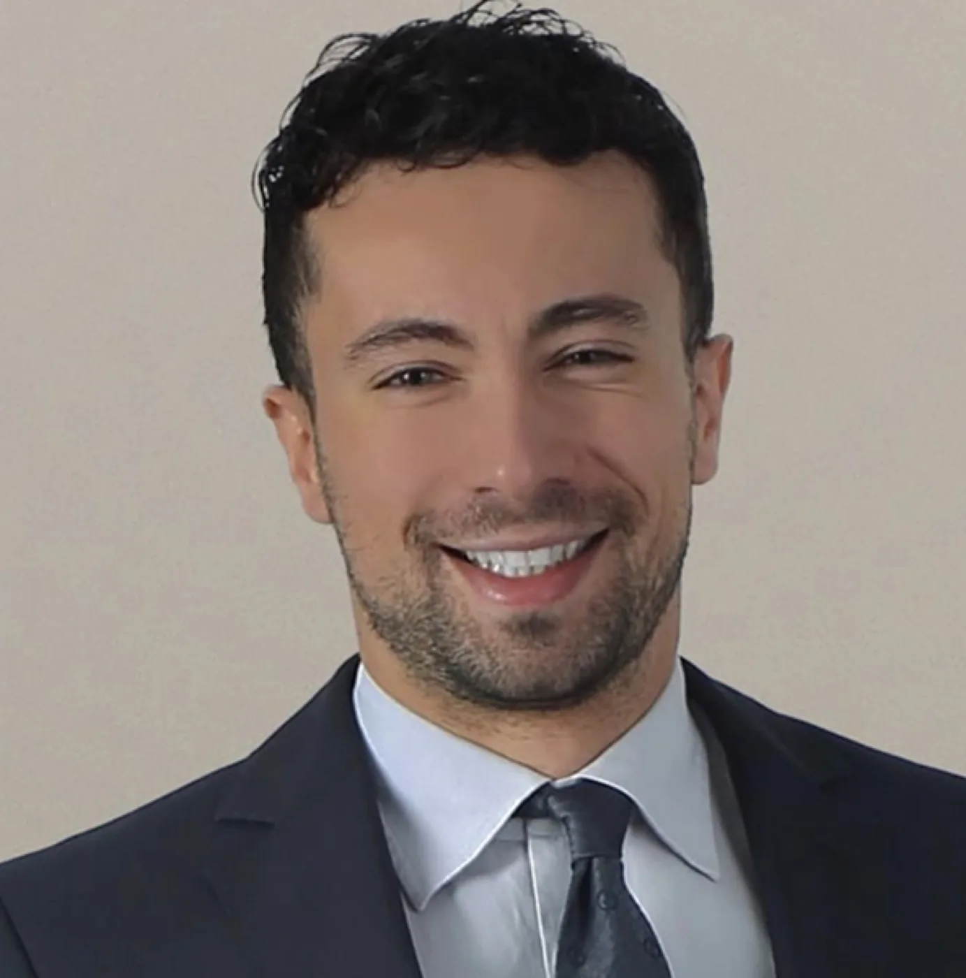 Professional headshot of Mr. Giuseppe Di Taranto, a man with short dark curly hair and a short beard, smiling broadly, wearing a dark navy suit jacket, light blue dress shirt, and a gray tie.