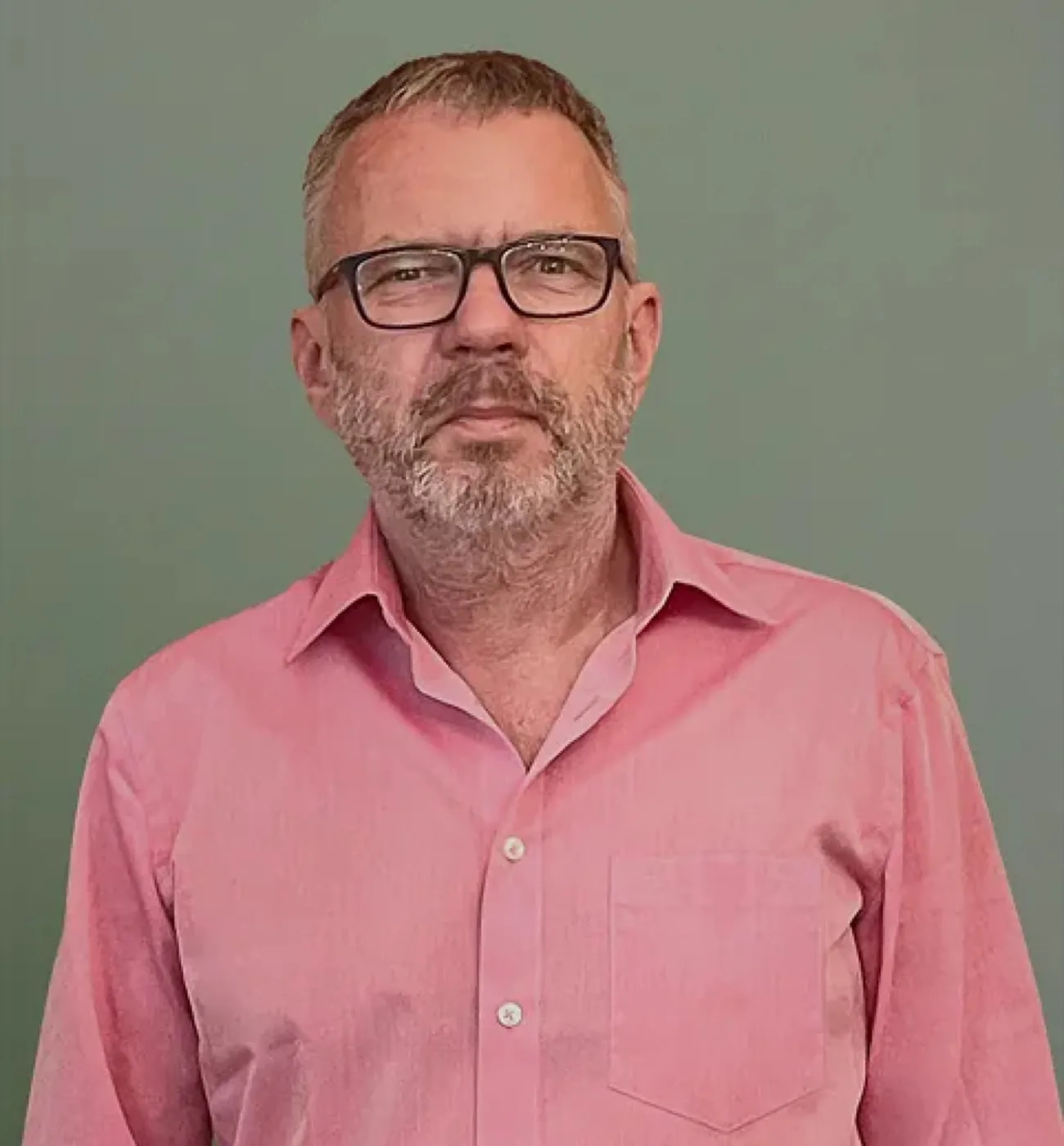 Professional headshot of Dr. Jörg Bohn, a middle-aged man with short salt-and-pepper hair and a gray beard, wearing black-rimmed rectangular glasses and a pink button-up shirt with a chest pocket, posed against a muted green background.