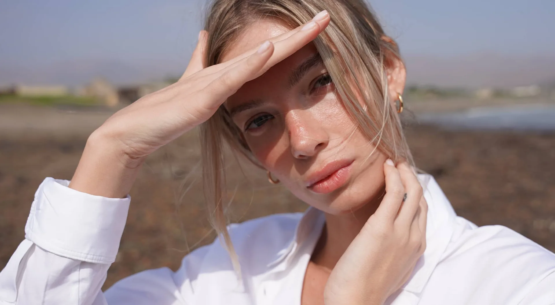 A young woman with blonde hair wearing a white button-up shirt, holding one hand to her forehead and the other to her neck, gazing at the camera with a serious expression. She is outdoors with a coastal landscape in the blurred background, in a hero image