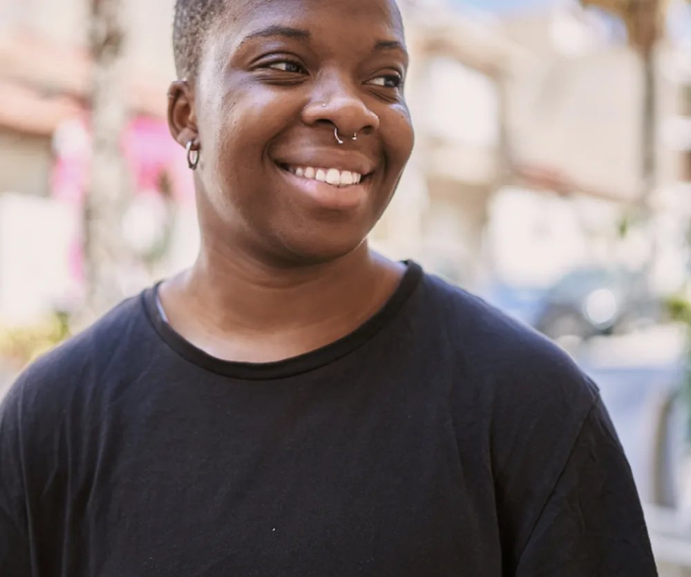 A person with short hair, a nose ring, and a small hoop earring, smiling warmly while wearing a black t-shirt, photographed outdoors with a blurred urban background.