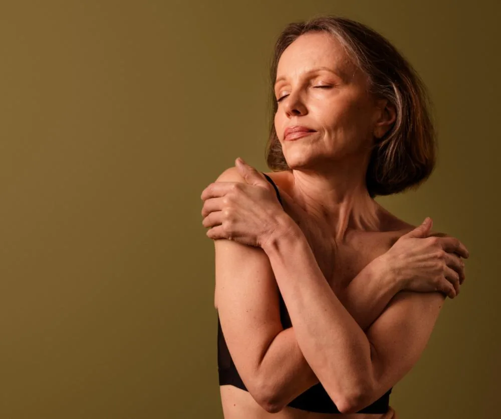 A middle-aged woman with short brown hair and closed eyes hugging herself gently across her shoulders, wearing a black bra, conveying a sense of calm self-acceptance — related to breast tissue preservation surgery.