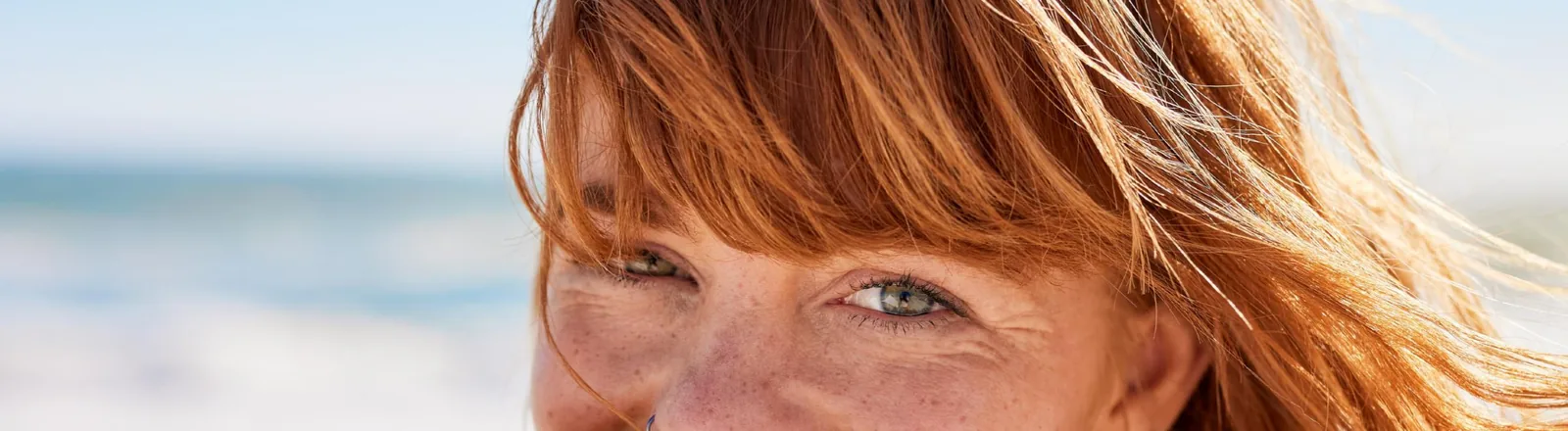 Close-up portrait of a middle-aged woman with auburn hair blowing in the wind, freckles, and green eyes, photographed outdoors near the ocean on a sunny day.