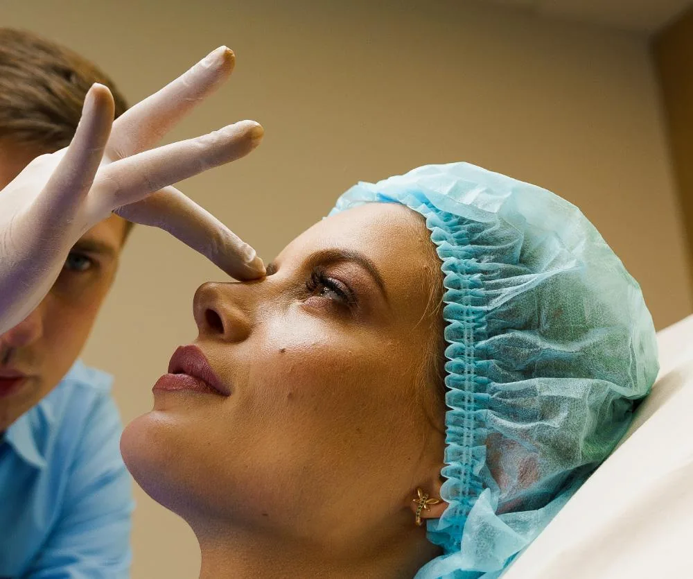 A female patient wearing a blue surgical cap lies reclined while a medical professional in latex gloves examines her nose area, likely in preparation for or following a septoplasty procedure.