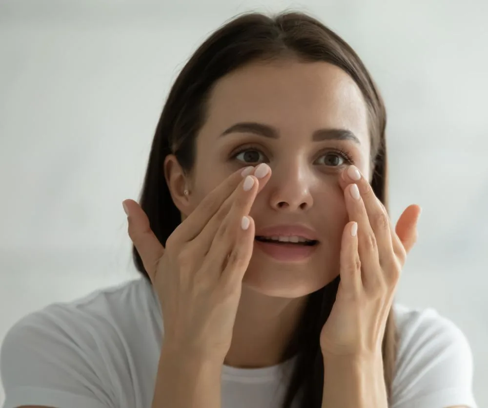 A young woman with dark hair and brown eyes gently applying cream or skincare product around her eyes with her fingertips, wearing a white t-shirt, illustrating post-blepharoplasty eye care.