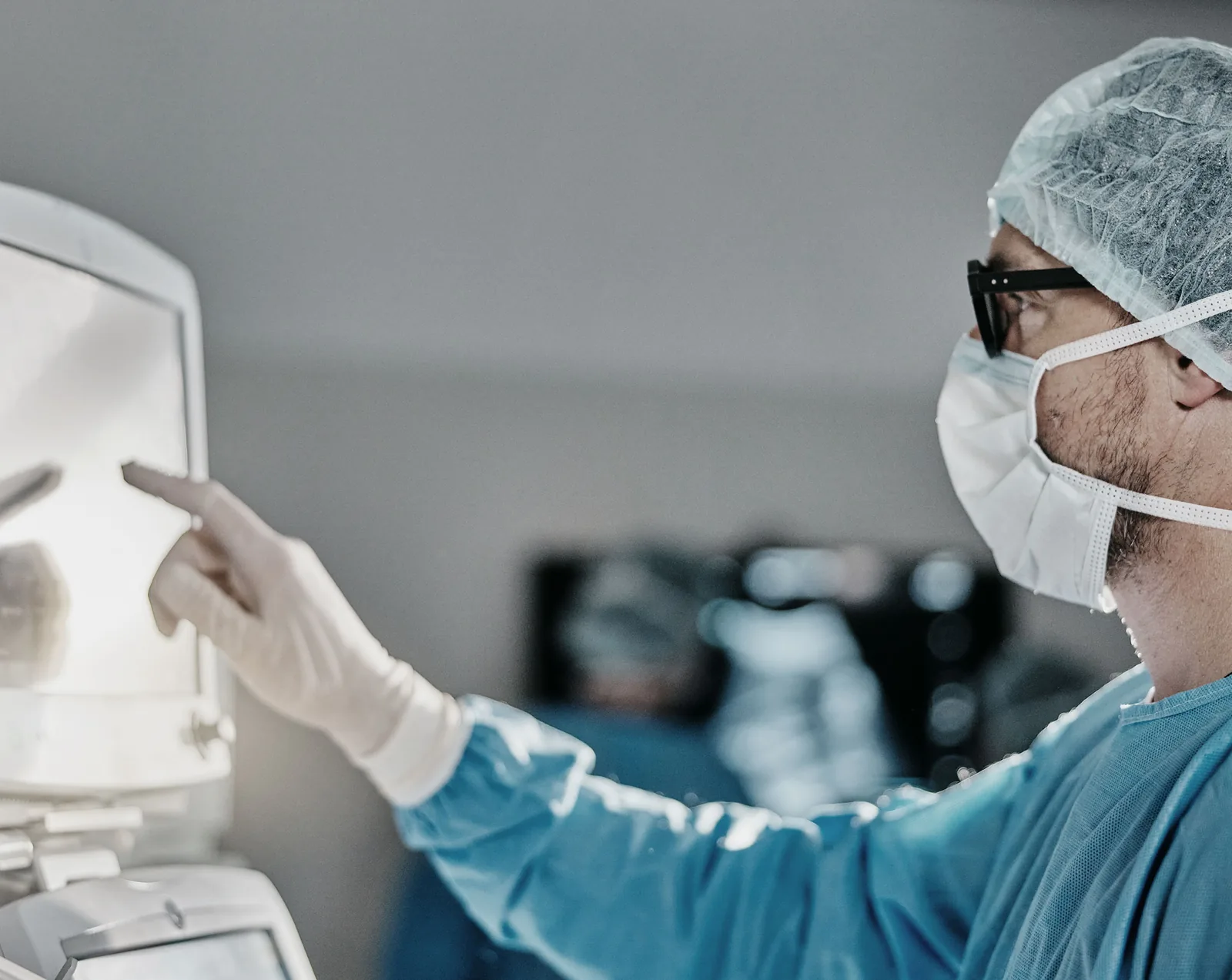 A male surgeon wearing blue scrubs, a teal surgical cap, white latex gloves, a white face mask, and black-framed glasses, pointing at a medical monitor screen in an operating room setting.