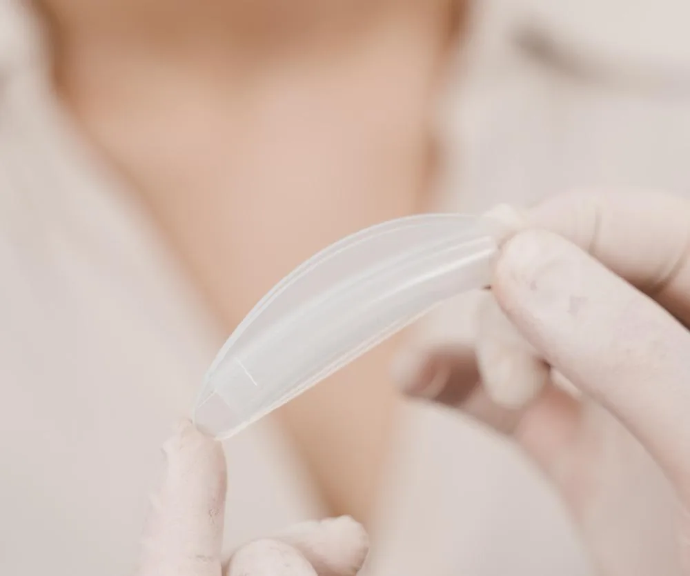 A gloved hand holding a clear, flexible silicone septoplasty nasal splint, with a blurred person wearing a light-colored garment visible in the background.
