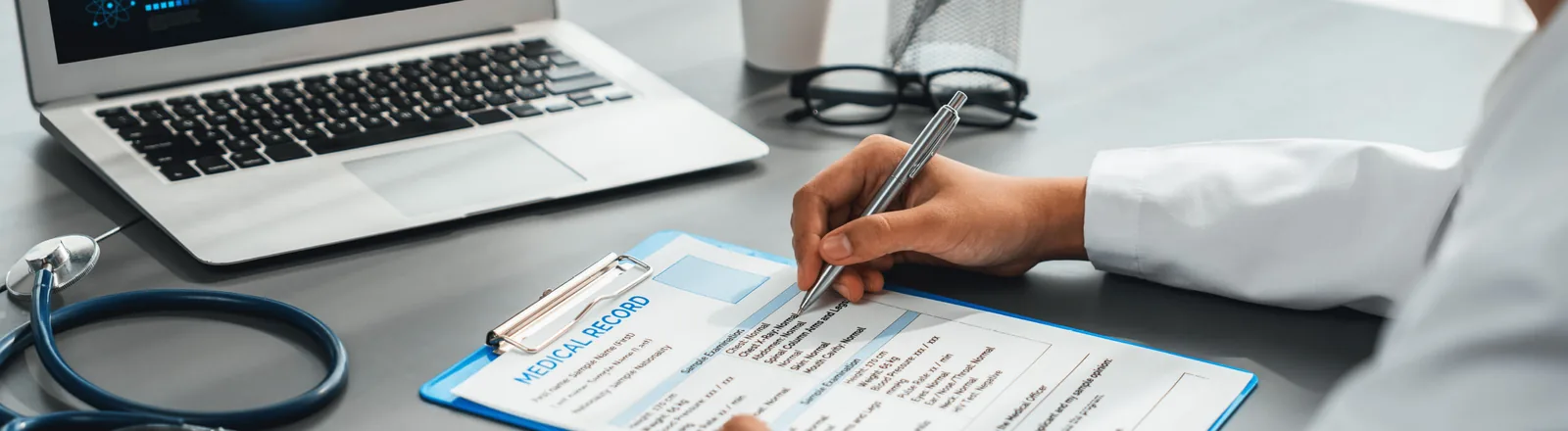 A doctor in a white coat writing on a medical record form attached to a blue clipboard, with a stethoscope, open laptop, eyeglasses, and a cup visible on the desk.