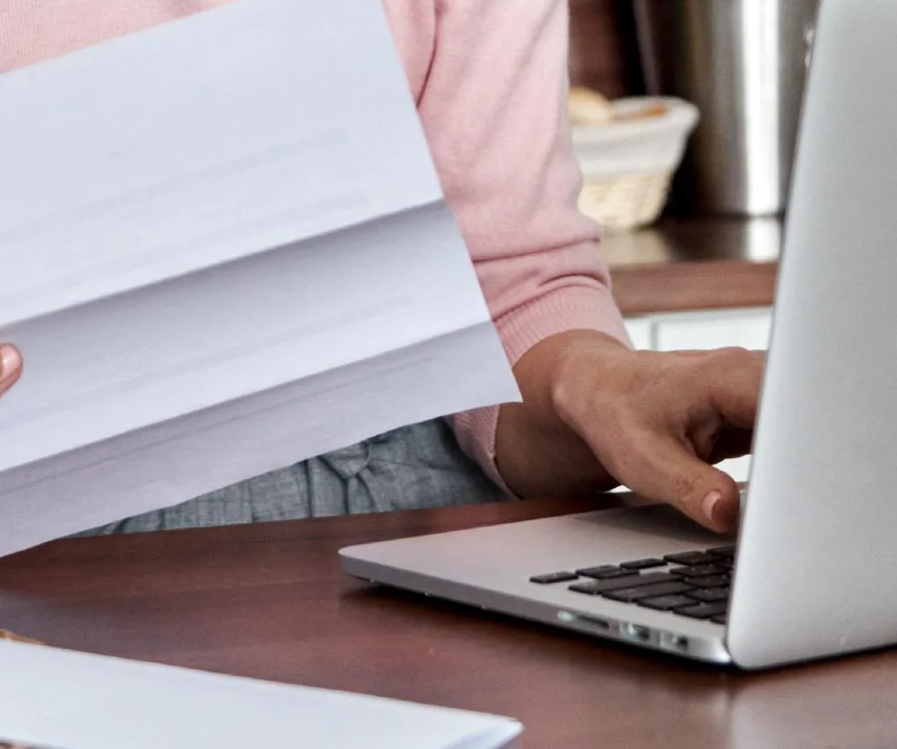 A person wearing a pink sweater holding a stack of papers in one hand while typing on a silver laptop with the other hand, seated at a wooden desk.