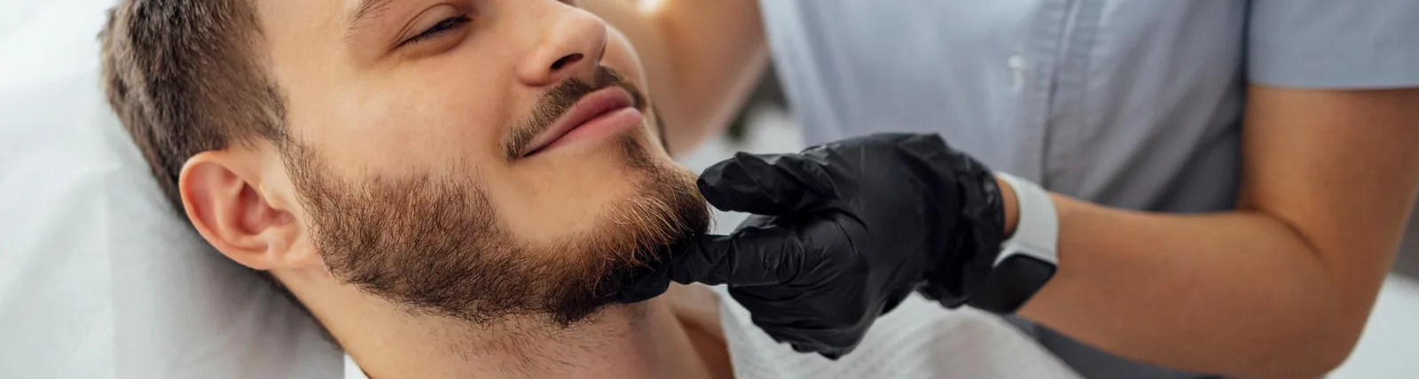 A smiling young man with a beard lying back while a gloved practitioner examines or touches his chin area, suggesting a Botox or cosmetic facial treatment consultation.