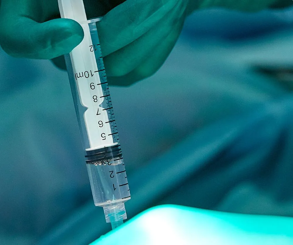 A gloved hand holding a 10ml medical syringe filled with local anaesthetic, used in a blepharoplasty (eyelid surgery) procedure, photographed under teal-toned surgical lighting.