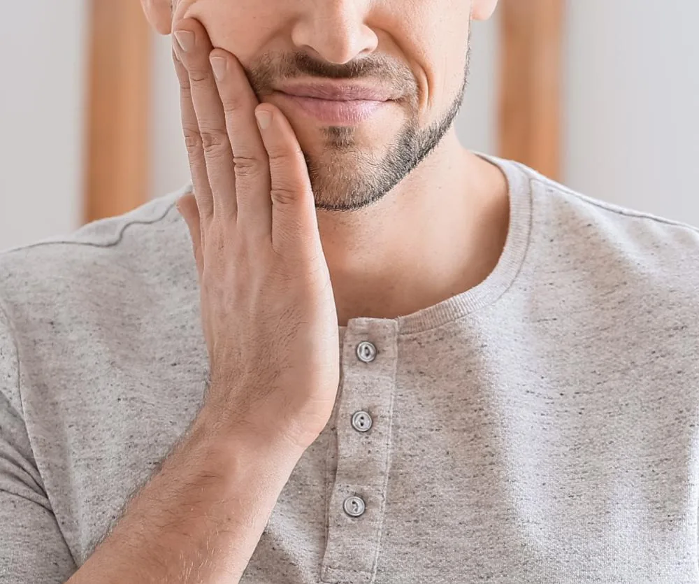 A man with a short beard holding his hand against his jaw and cheek, grimacing in apparent tooth or jaw pain, wearing a light gray henley shirt.