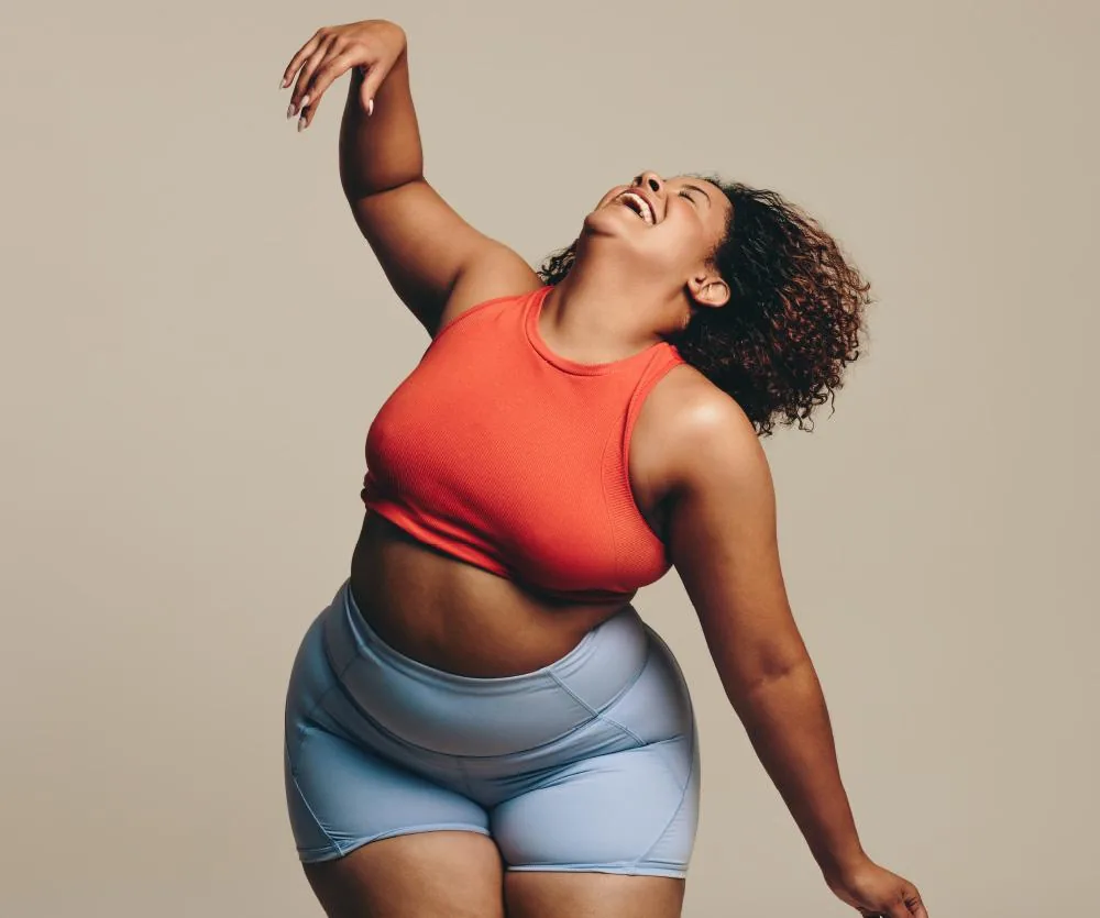A plus-size woman joyfully posing in a red sports crop top and light blue athletic shorts, laughing with one arm raised, conveying body confidence and positivity.