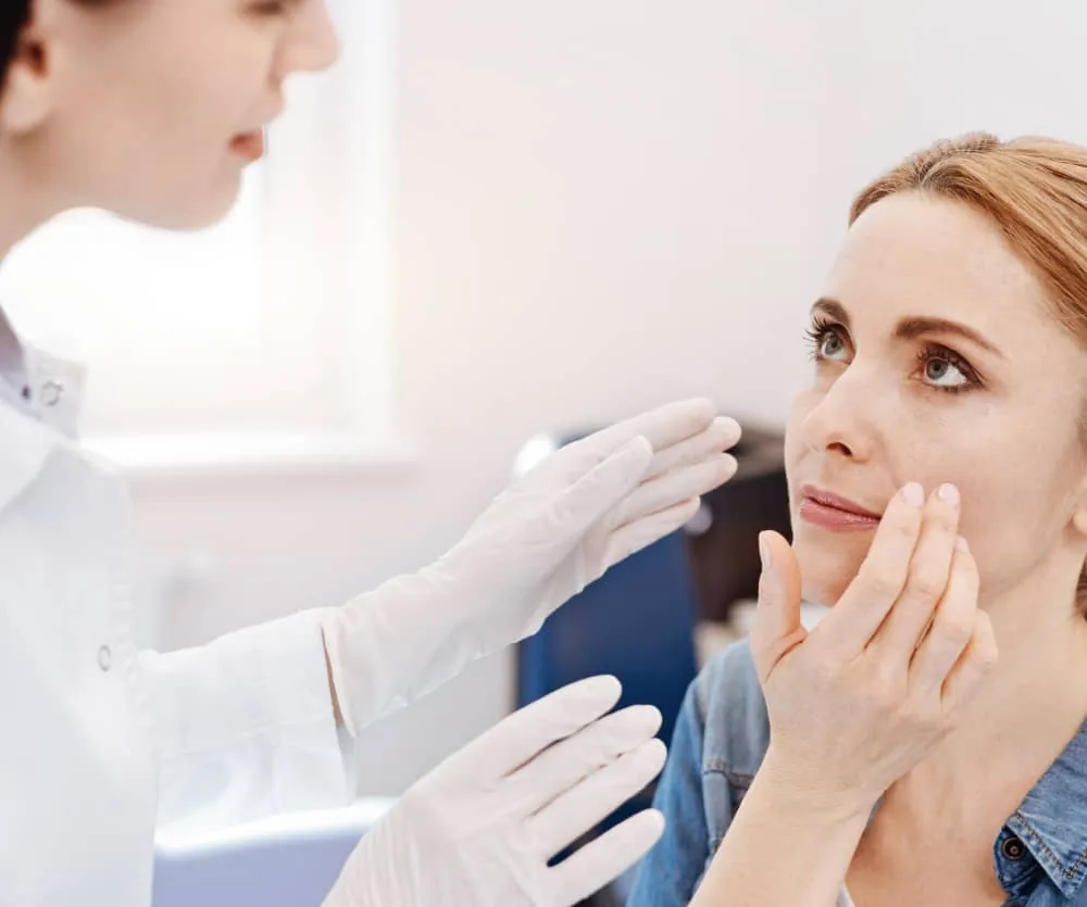 A medical professional wearing white latex gloves gently examining and touching the cheeks of a blonde woman's face during a non-surgical buccal fat removal consultation in a clinical setting.