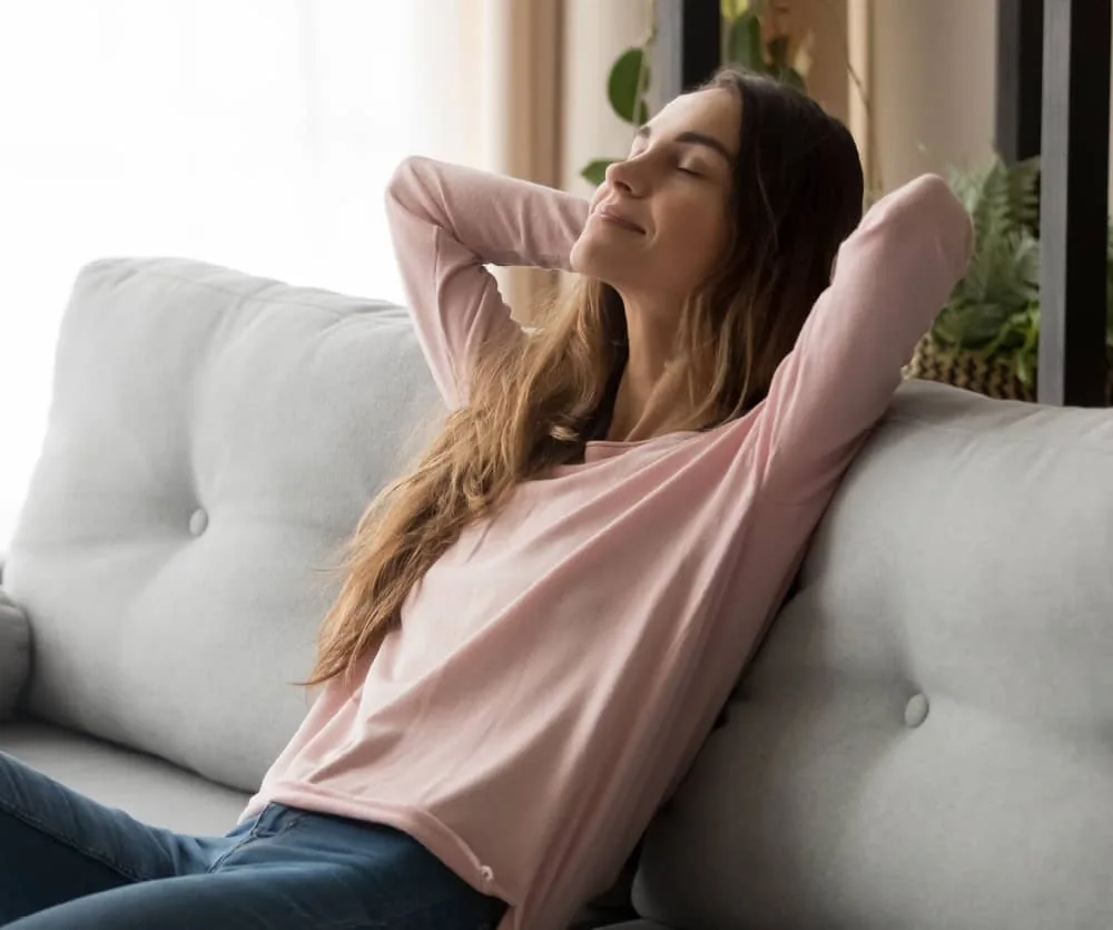 A young woman with long brown hair relaxing on a light gray sofa, leaning back with her eyes closed and hands clasped behind her head, wearing a pink long-sleeve top and blue jeans, with a plant and bright window visible in the background.